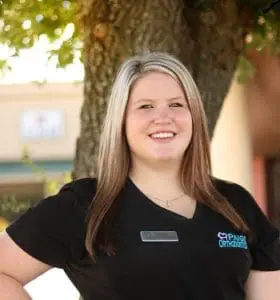 A woman in a black shirt stands smiling, showing off her braces, as she poses outdoors in front of a tree.