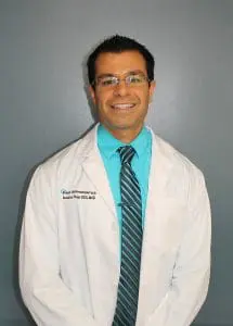 A person in a white lab coat and glasses, possibly an orthodontist from Porter, TX, smiles at the camera. They wear a turquoise shirt and striped tie, standing confidently against a gray background.