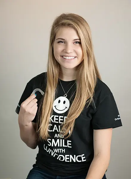 A woman with long hair smiles, proudly holding her Invisalign at Paige Orthodontics in Kingwood, TX. She wears a black T-shirt with a smiley face and the text: "KEEP CALM AND SMILE WITH CONFIDENCE," showcasing her bright attitude.