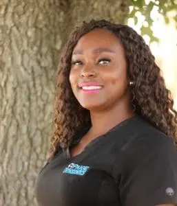 A person with long curly hair and a black shirt smiles, standing outdoors near a tree trunk in Porter, TX.