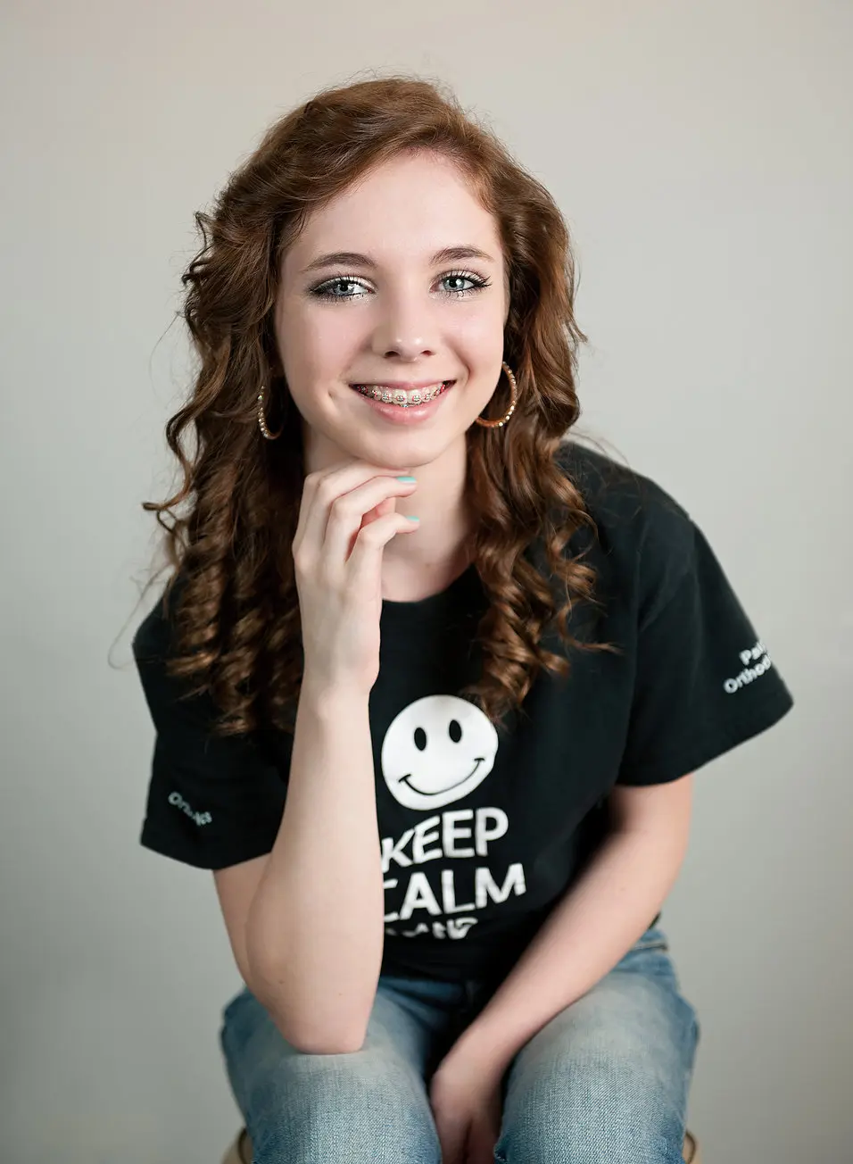 A young person from Paige Orthodontics in Kingwood TX, with curly hair, metal braces, hoop earrings, and a "Keep Calm" T-shirt, smiles while resting their chin on their hand against a plain background.