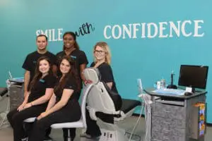 In the Paige Orthodontics office in Kingwood TX, five dental professionals wearing black uniforms pose confidently against a turquoise wall featuring the phrase "Smile with Confidence.