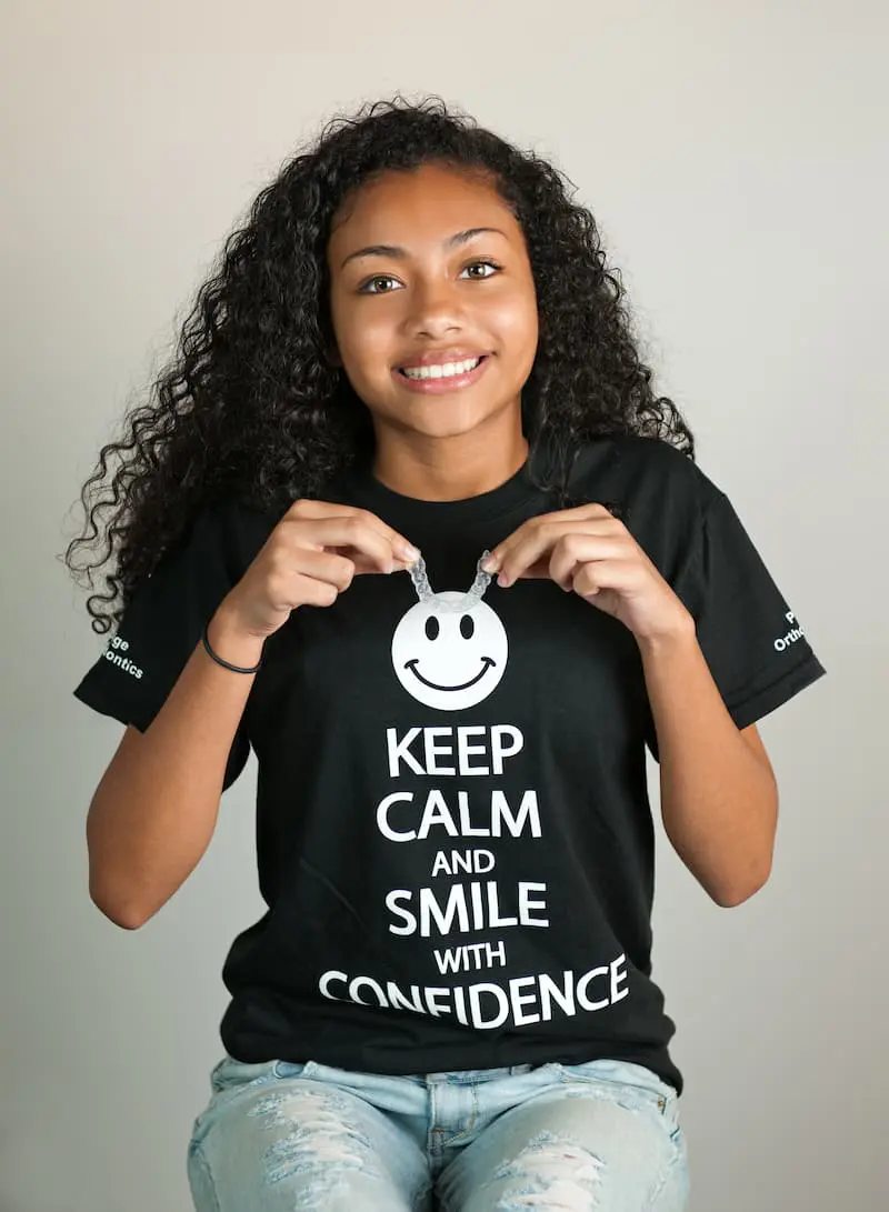 In Kingwood TX, a teen girl with curly hair smiles while holding Invisalign clear aligners. Her black t-shirt, "Keep Calm and Smile with Confidence," showcases her choice of Paige Orthodontics for a perfect smile.