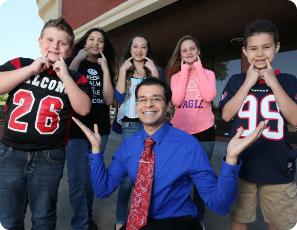 Dr. Sebastian Paige in a blue shirt and red tie, as an orthodontist from Kingwood, poses in front of six smiling children standing outside a TX building.