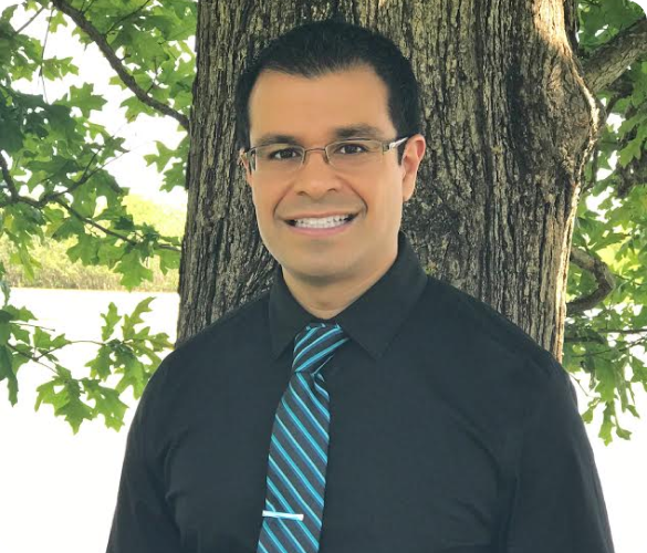 Dr. Sebastian Paige in glasses and a black shirt with a blue striped tie, perhaps an orthodontist in Kingwood, TX, stands outdoors in front of a tree.