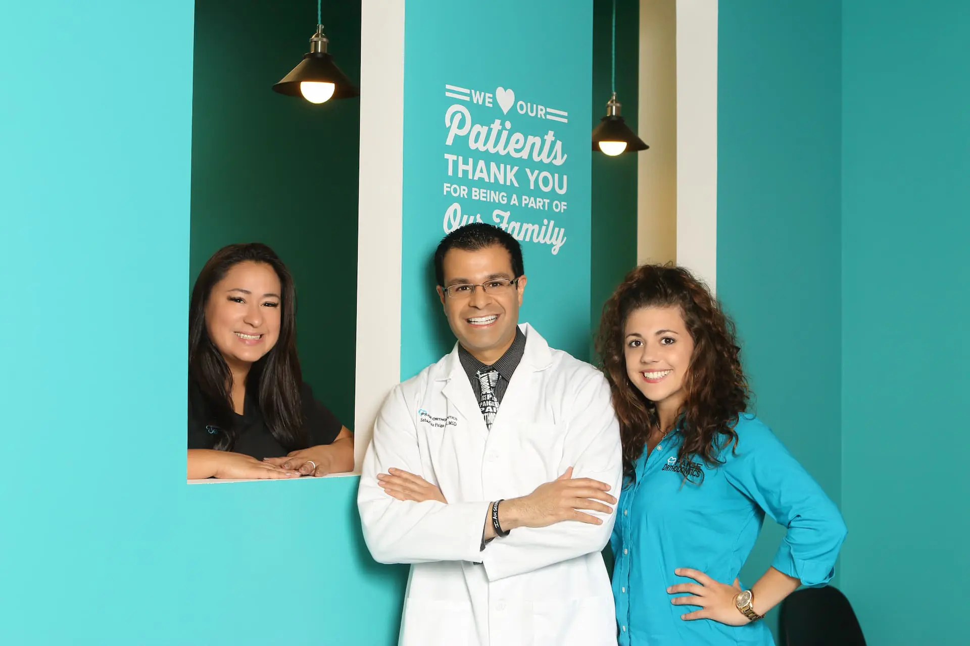 In a teal room in Kingwood TX, three people smile: one in a white coat and two in blue tops. A wall sign thanks patients for their support. At Paige Orthodontics, Dr. Sebastian Paige values their smiles deeply.