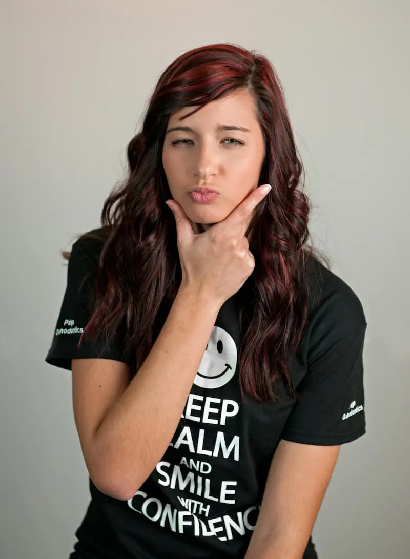 A person with long brown hair in Kingwood TX poses confidently with a hand on their chin, wearing a black "Keep Calm and Smile with Confidence" T-shirt, proudly showing off their Paige Orthodontics smile.