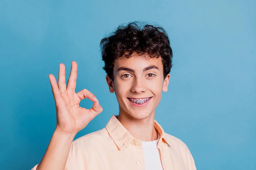 A teen from Kingwood TX with curly hair, braces, and a slight overbite makes an "OK" hand gesture against a blue background.