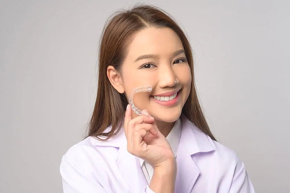 Against a plain background, a person in a white coat confidently smiles while holding a clear Invisalign aligner in Kingwood TX.