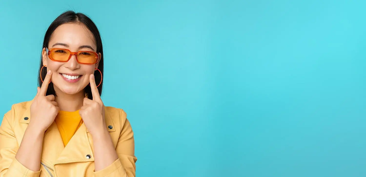 Against a blue background, a woman in a yellow jacket and orange sunglasses smiles, pointing at her cheeks to highlight the benefits of the Herbst Appliance. Such orthodontic advancements can be explored further in Kingwood TX.