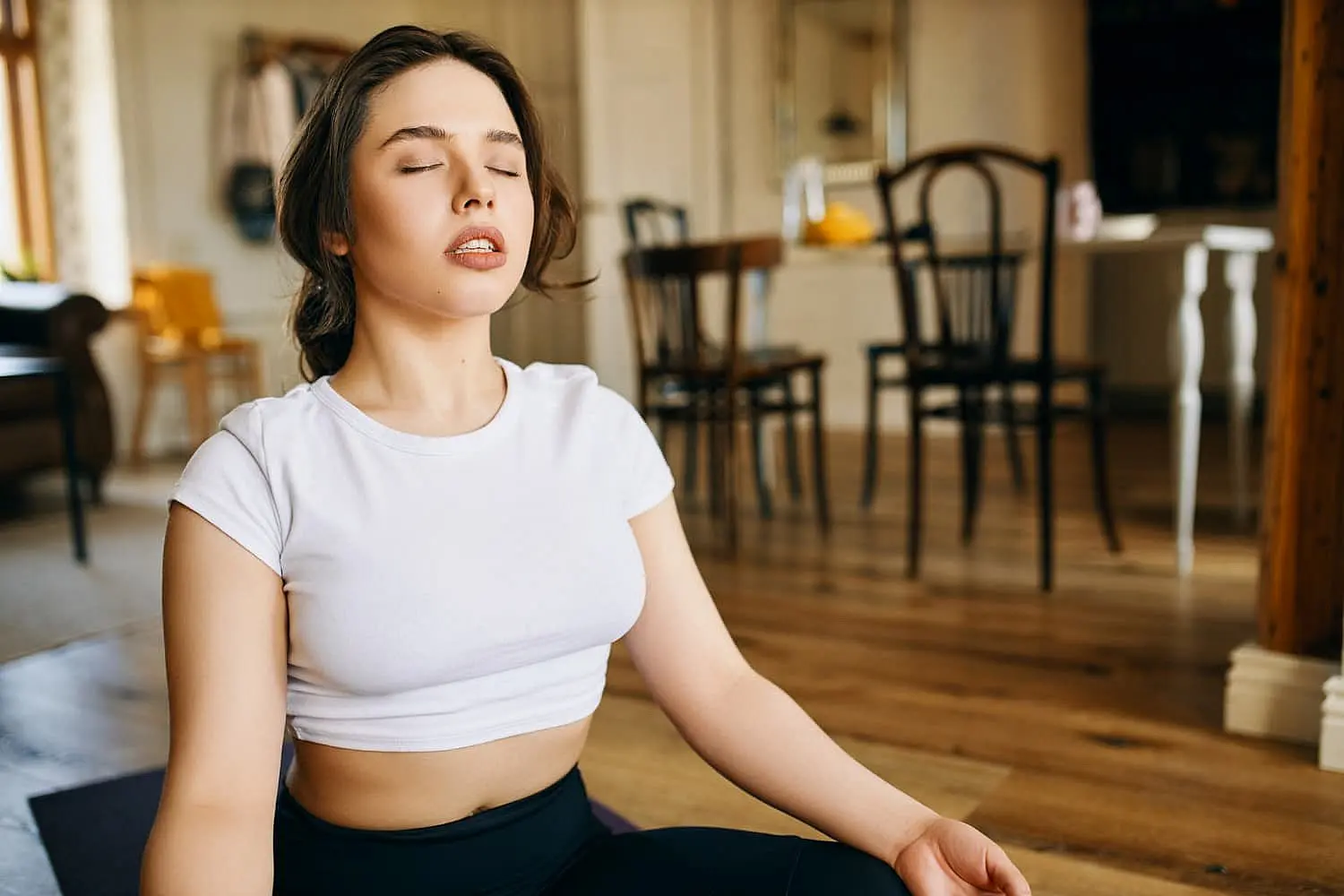 In a tranquil room in Kingwood TX, a person meditates on a yoga mat, wearing a white shirt. Eyes gently closed, they focus on transitioning from mouth breathing to soothing nasal breaths amidst chairs and a table.