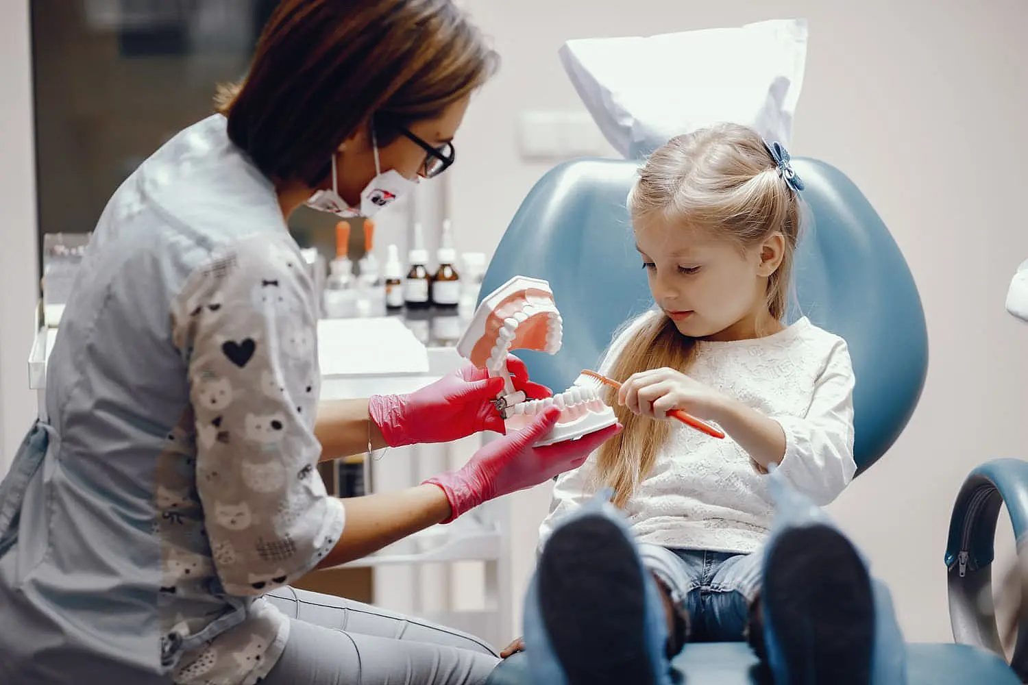a dentist demonstrates tooth brushing to a young girl seated in a dental chair with a toothbrush at Paige Orthodontics in Kingwood, TX