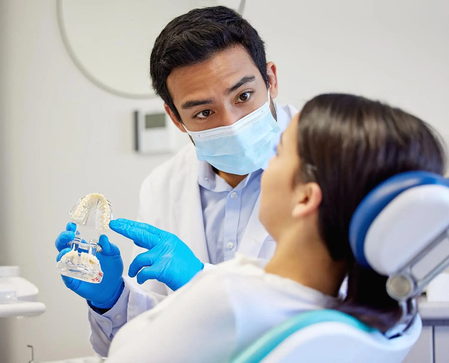 In Kingwood TX, a dentist, equipped with a mask and gloves, explains dental procedures to a patient using a tooth model and discusses the use of a palatal expander.