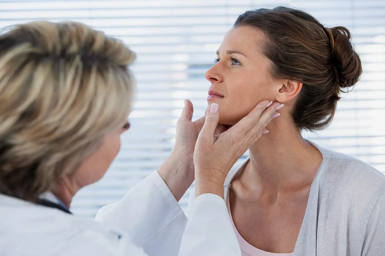 In a medical office in Kingwood TX, with blinds in the background, a doctor examines a woman's face and neck to assess whether jaw surgery is necessary.