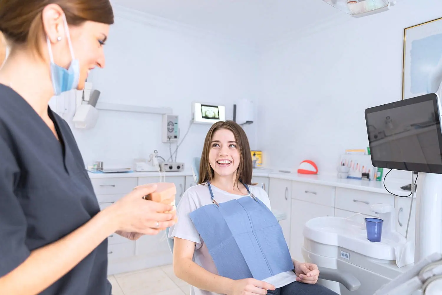 In Kingwood TX, a woman with metal braces sits in a dental chair, smiling at a dentist holding a cup; dental equipment and a monitor are visible in the background.