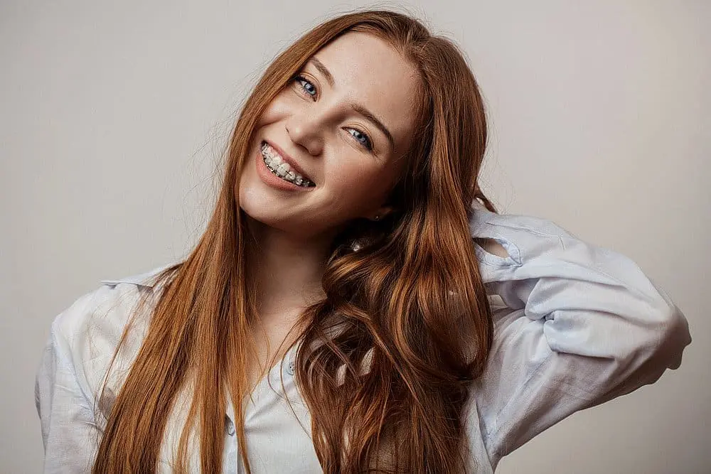 a person with long red hair, braces, and a light-colored shirt smiles at Paige Orthodontics in Kingwood, TX