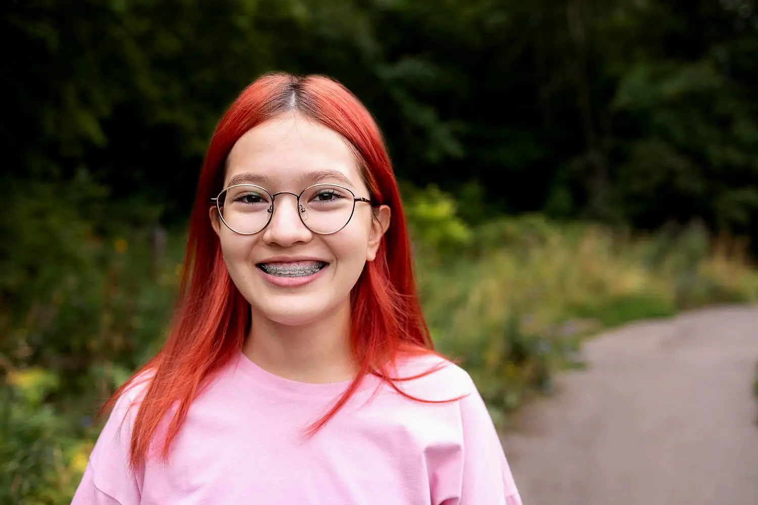 In a lush, green park in Kingwood TX, a red-haired person with glasses and an endearing underbite wears a pink shirt and smiles warmly on the path.