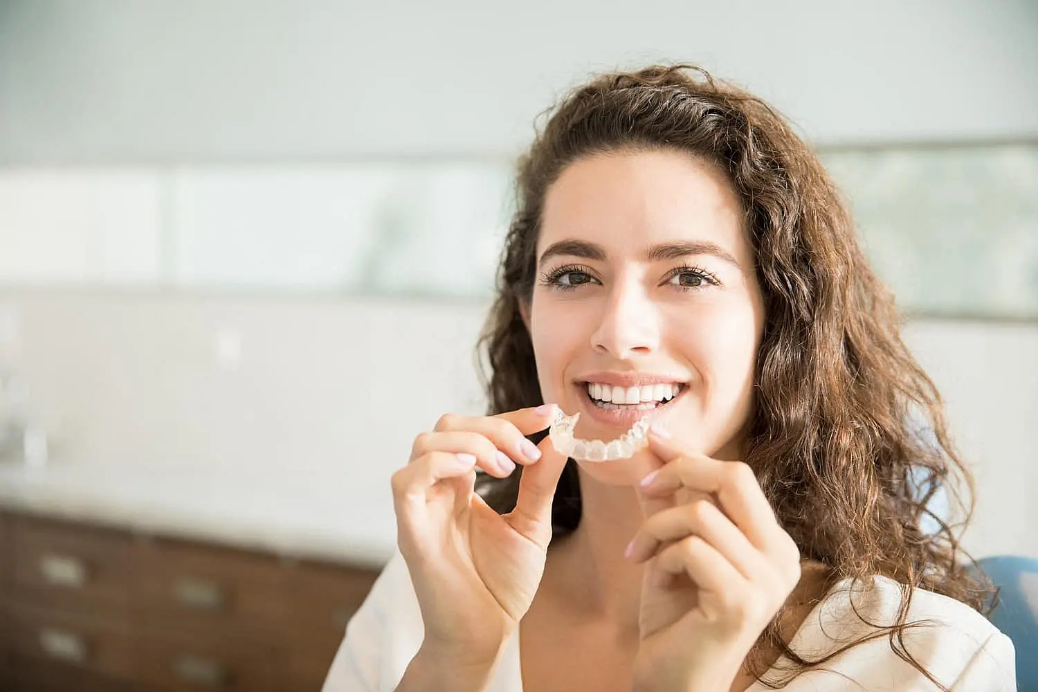 In Kingwood TX, a woman with long curly hair smiles while holding a clear dental aligner near her mouth.