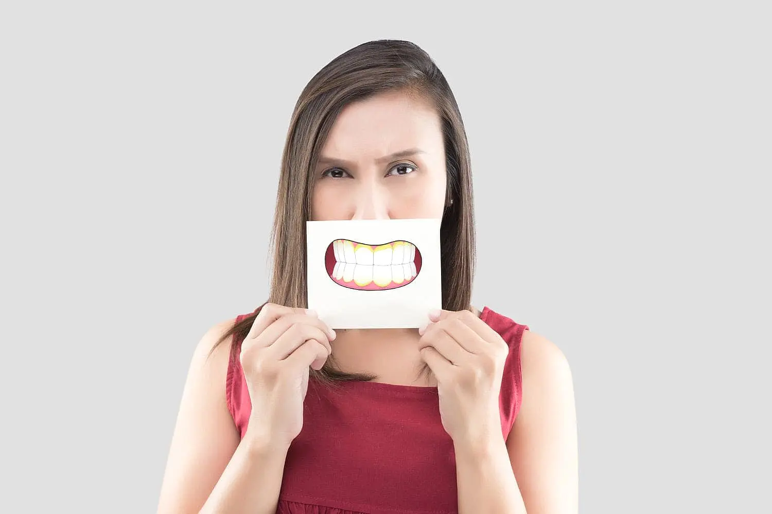 In Kingwood TX, a woman in a red top playfully holds a paper with a cartoonish smiling mouth, suggesting an exaggerated underbite against a plain background.