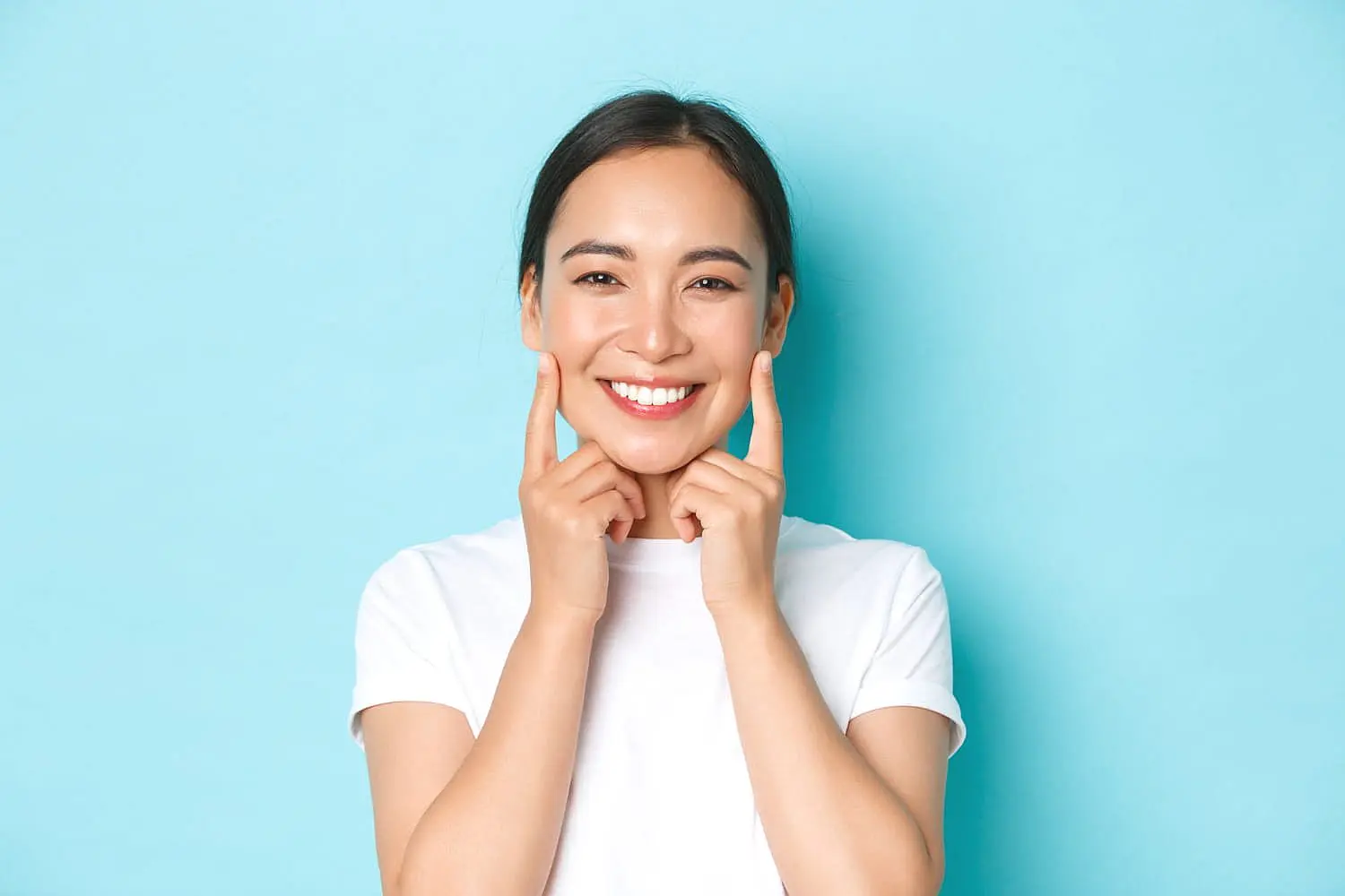Against a blue background, a person in a white t-shirt with an endearing overbite smiles and points to their cheeks in Kingwood TX.