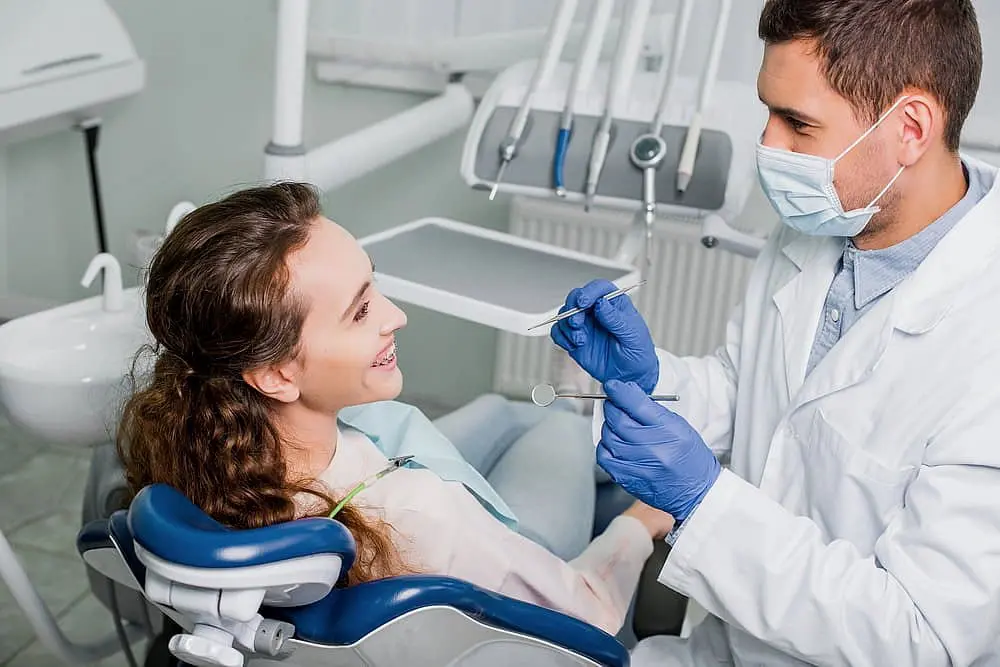In Kingwood TX, a dentist in mask and gloves examines a smiling woman with metal braces, surrounded by dental tools and equipment.