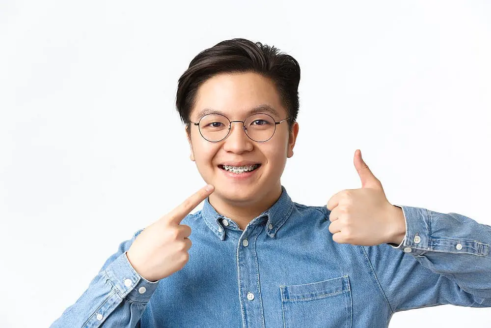 Beaming in Kingwood TX, a person in a denim shirt and glasses proudly points at their adult braces with one hand and gives a thumbs-up with the other against a plain backdrop.