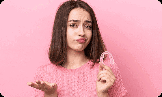 Against a pink background, a woman in a pink sweater with long brown hair looks puzzled while holding an Invisalign retainer in Kingwood TX.