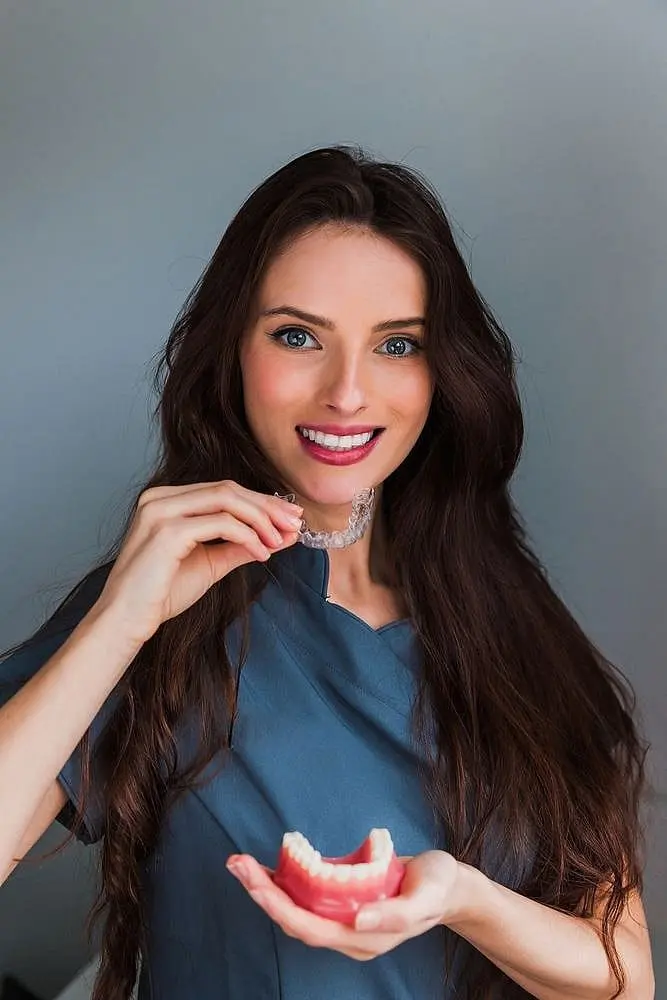 Against a gray backdrop, a woman in a blue shirt smiles warmly while displaying an Invisalign dental aligner and a model of teeth in Kingwood TX.
