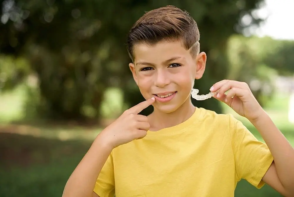 A boy in a yellow shirt smiles and points to his teeth, holding a clear dental aligner outside in Kingwood TX.