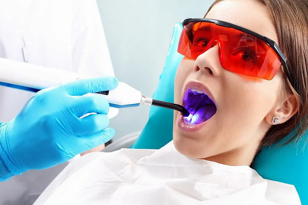 At Paige Orthodontics in Kingwood TX, a dentist uses a dental curing light with precision on a patient wearing red protective glasses. This clinic is also known for its expertise in Invisalign vs Braces to help you achieve the perfect smile.