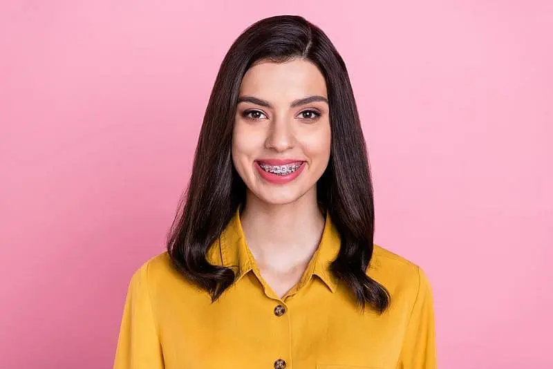 A person with long dark hair and braces smiles in a yellow shirt set at Paige Orthodontics in Kingwood TX.
