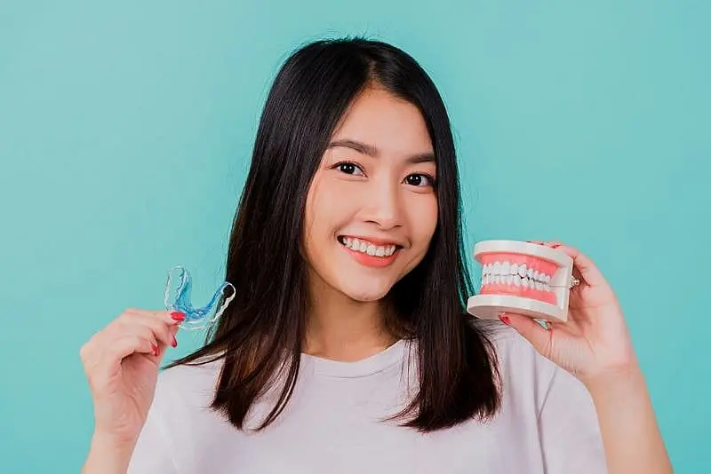a smiling person holds both a dental retainer and a model set of teeth at Paige Orthodontics in Kingwood, TX