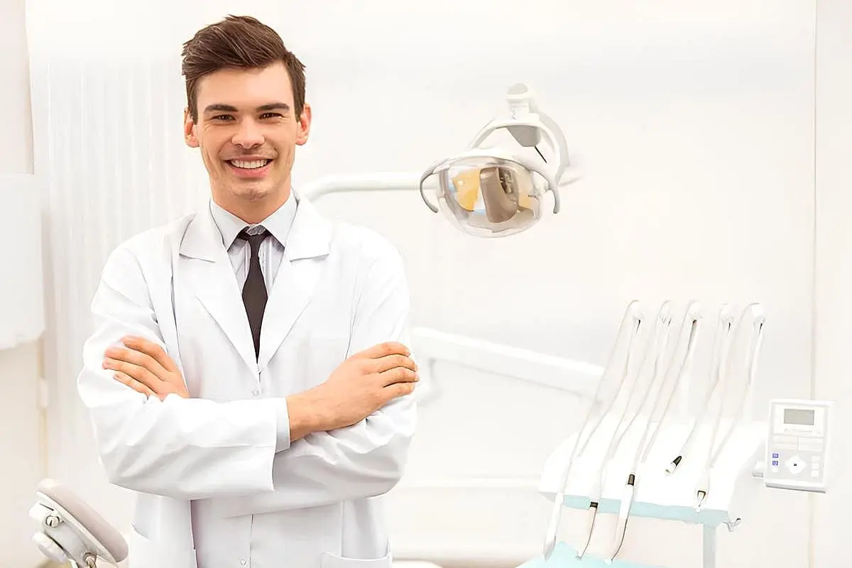 In Kingwood TX, a dentist in a white coat stands confidently with arms crossed in a dental office. Visible dental equipment showcases expertise in procedures like jaw surgery.