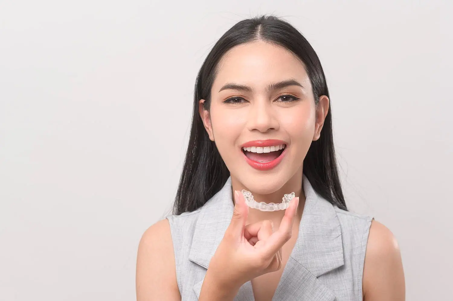 a woman with long dark hair smiles, holding a clear dental retainer near her mouth at Paige Orthodontics in Kingwood, TX
