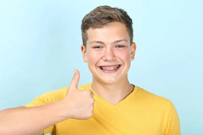 Against a light blue background, a teen boy from Kingwood TX with metal braces and a yellow shirt gives a thumbs-up.