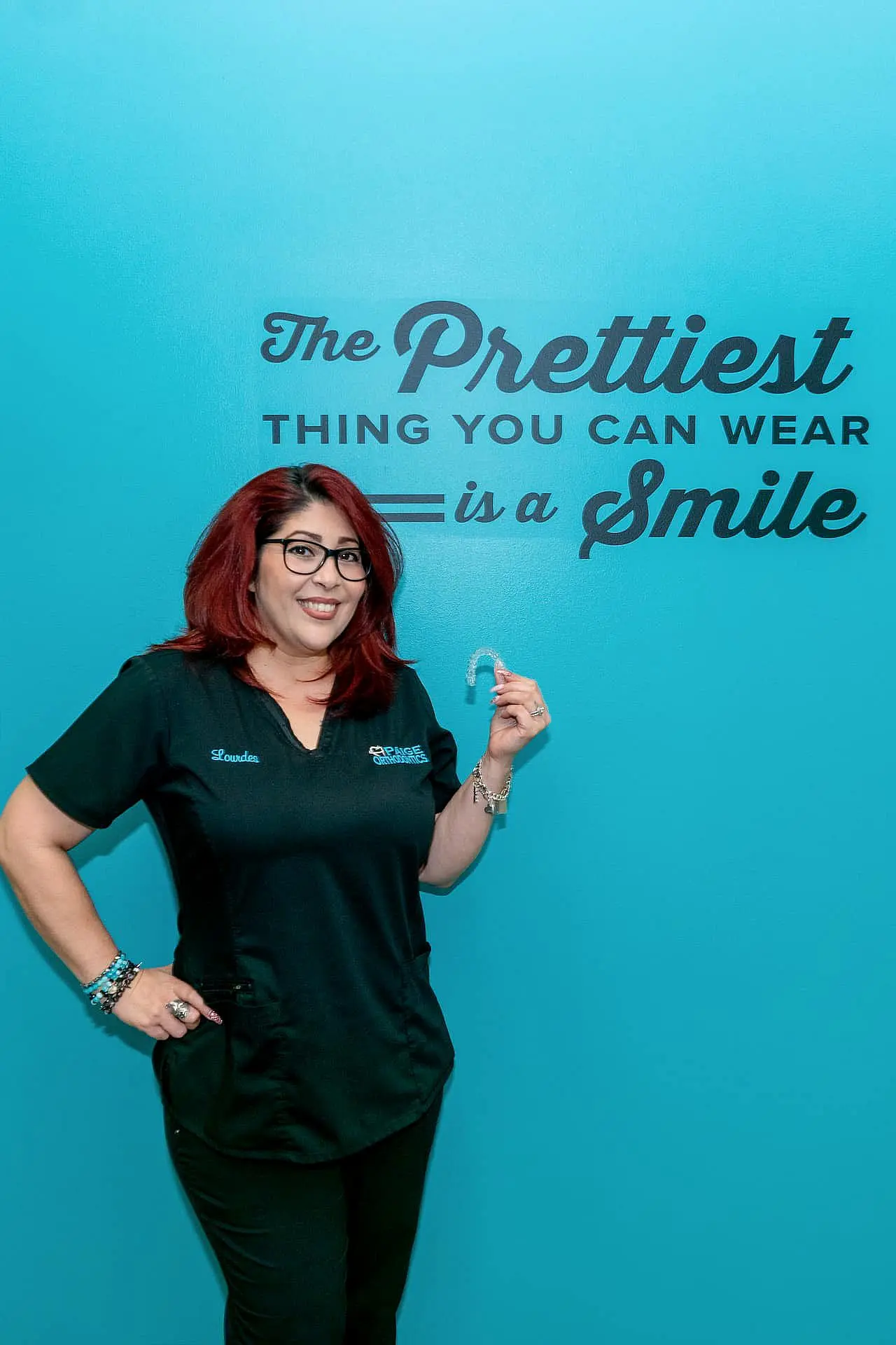 In Kingwood TX, a red-haired woman smiles against a teal wall while holding a dental aligner, supported by orthodontic insurance. The wall features the quote, "The prettiest thing you can wear is a smile.