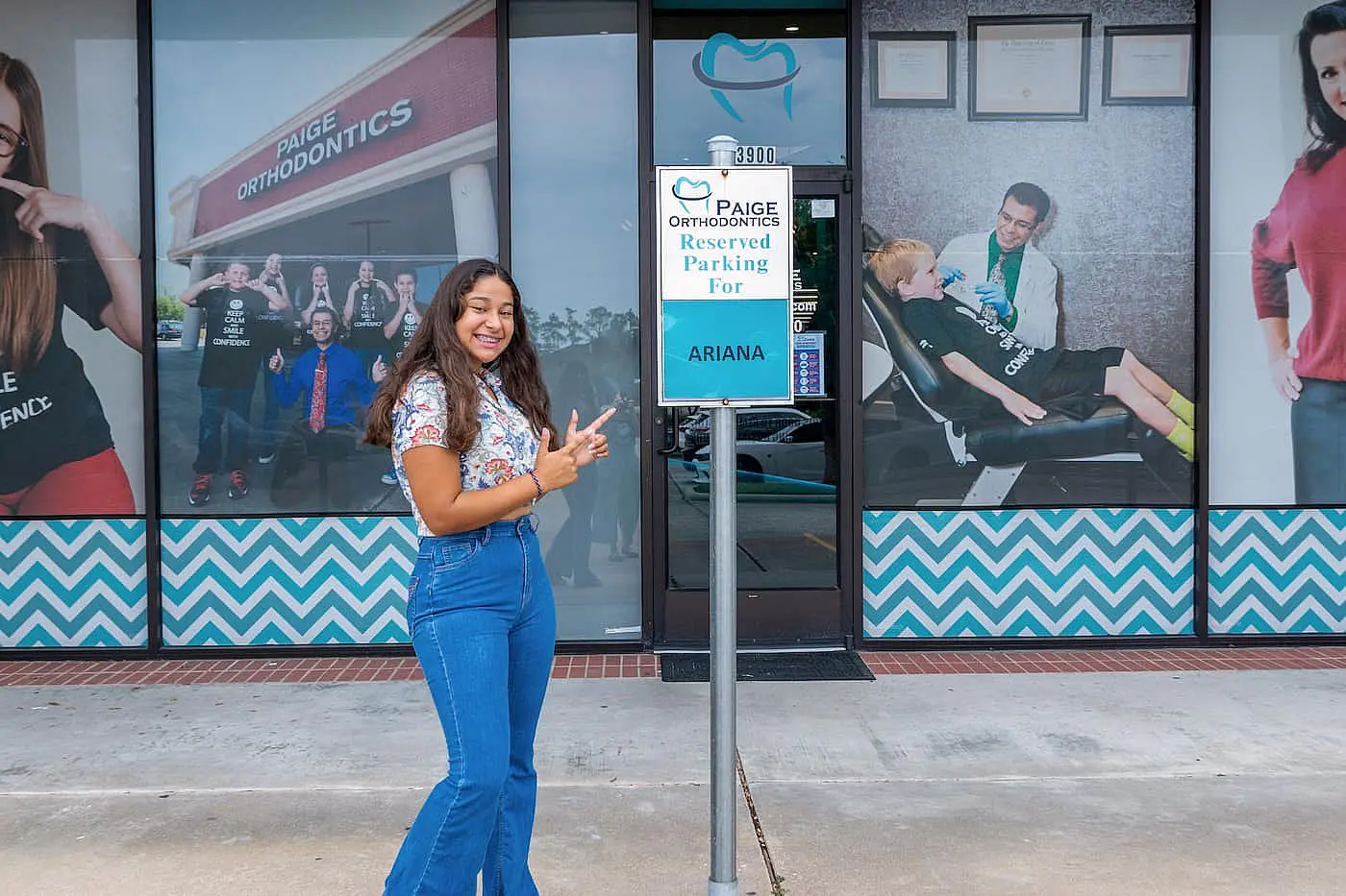 Next to the "Ariana" reserved parking sign outside an orthodontics office in Kingwood TX, a smiling person stands as posters of metal braces decorate the windows.