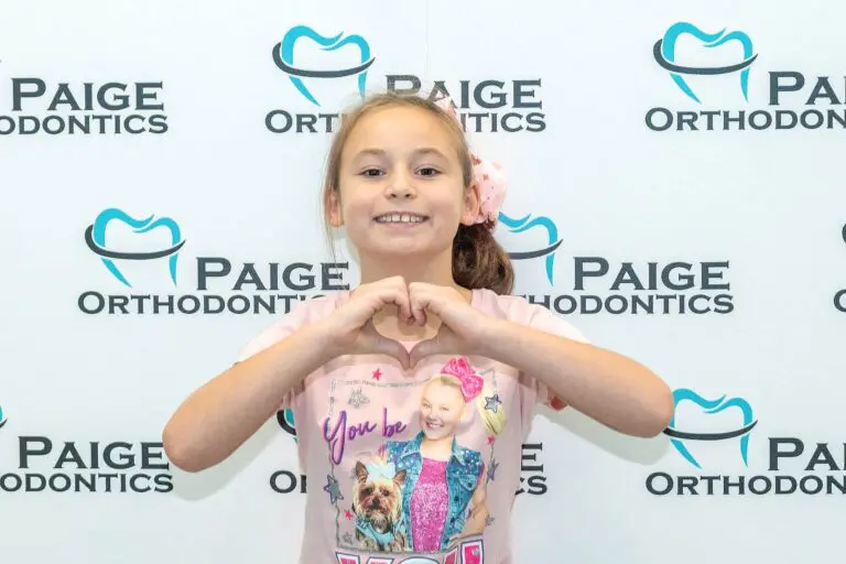 Against a "Paige Orthodontics" backdrop, a girl in a dog and girl print shirt forms a heart with her hands. Her smile reflects the confidence from having orthodontic insurance in Kingwood TX.