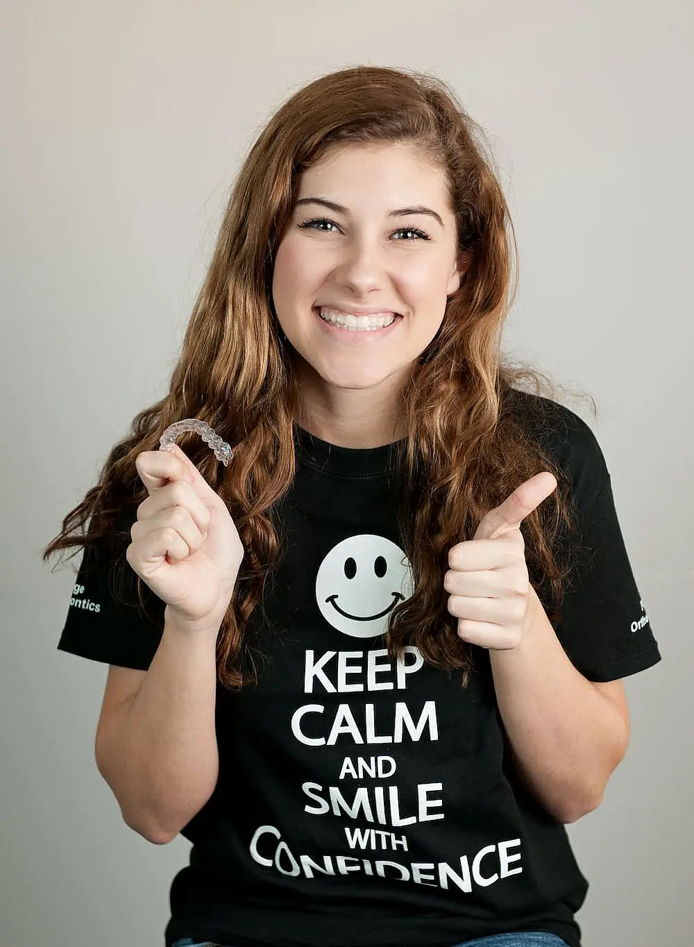 In Kingwood TX, a person smiles while holding a dental retainer and giving a thumbs-up, dressed in a shirt featuring a smiley face and motivational text.