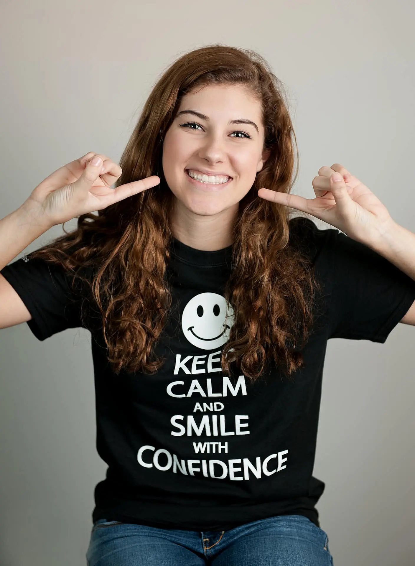 In Kingwood TX, a woman confidently smiles and points to her cheeks, wearing a black T-shirt with a smiley face and the message, "Keep Calm and Smile with Confidence," reflecting her Invisalign vs. braces journey.