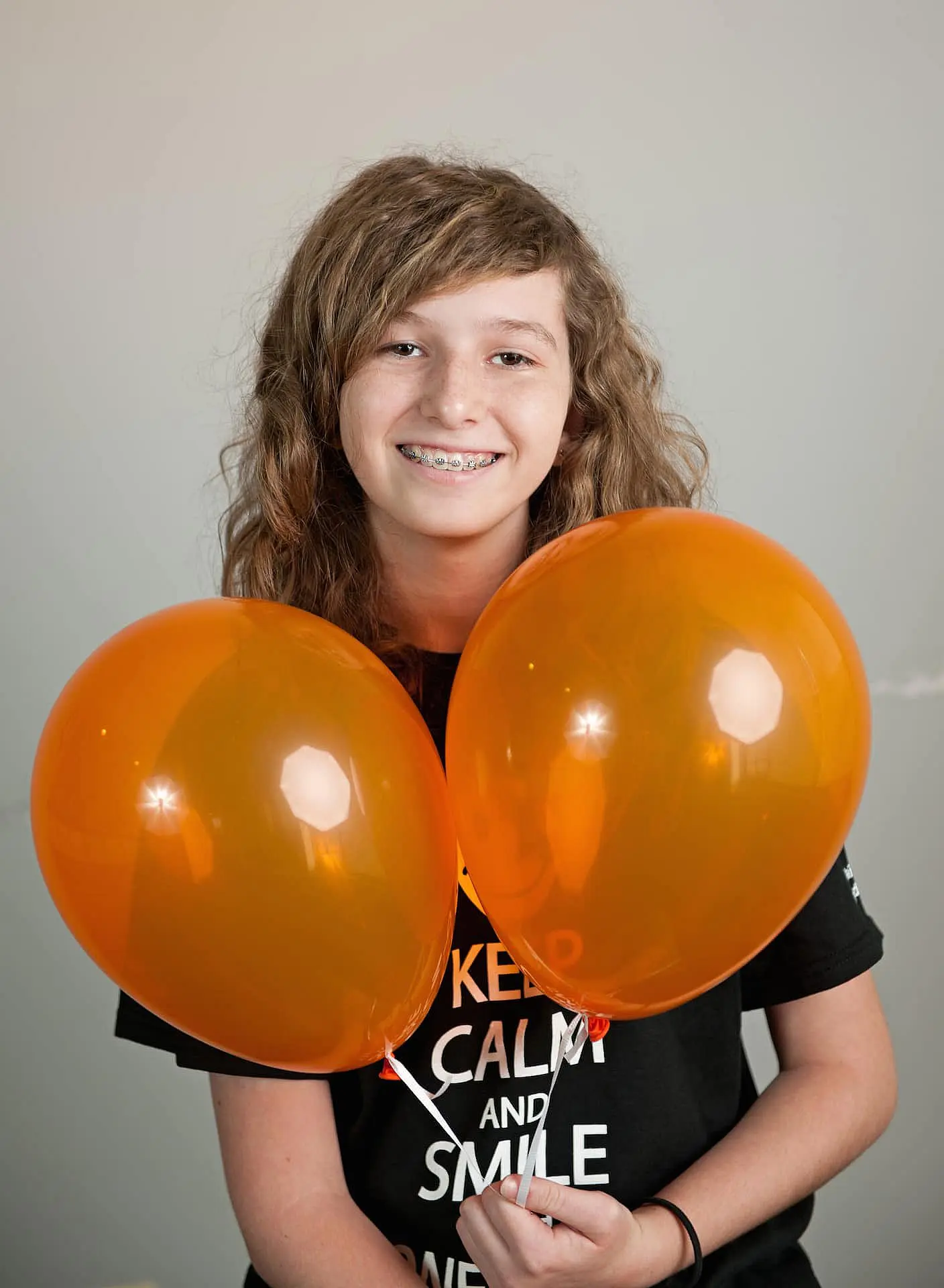 In Kingwood TX, a smiling individual with long hair and braces holds two orange balloons while wearing a black T-shirt.