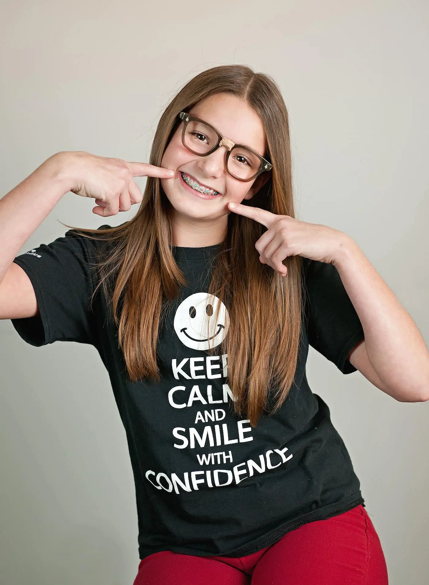 In Kingwood TX, a girl with long hair and glasses smiles and points to her braces. She wears a black shirt reading "Keep Calm and Smile with Confidence" paired with red pants.