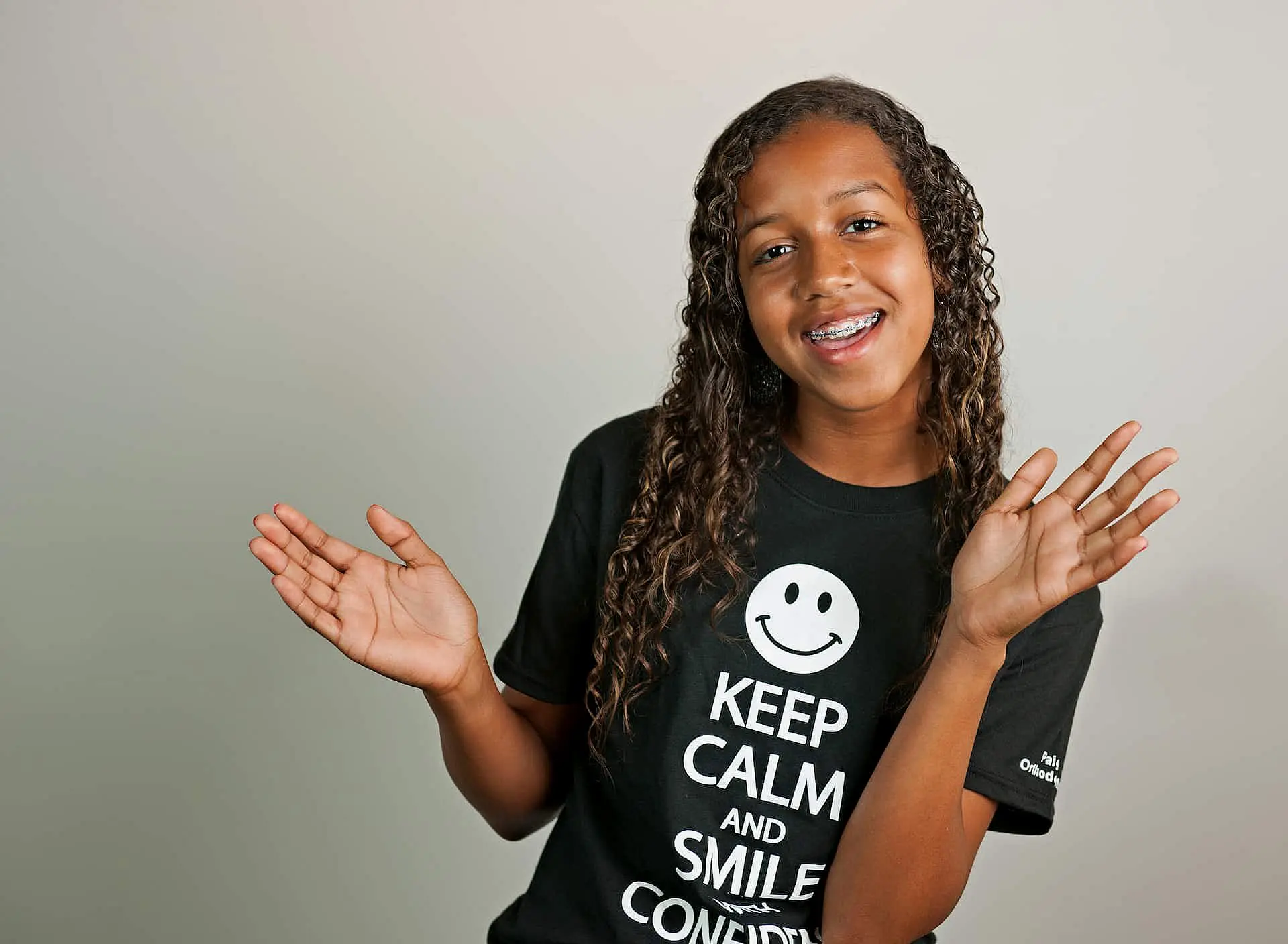 In Kingwood TX, a smiling person with long curly hair and braces playfully raises their hands, wearing a "Keep Calm and Smile" T-shirt against a plain background.