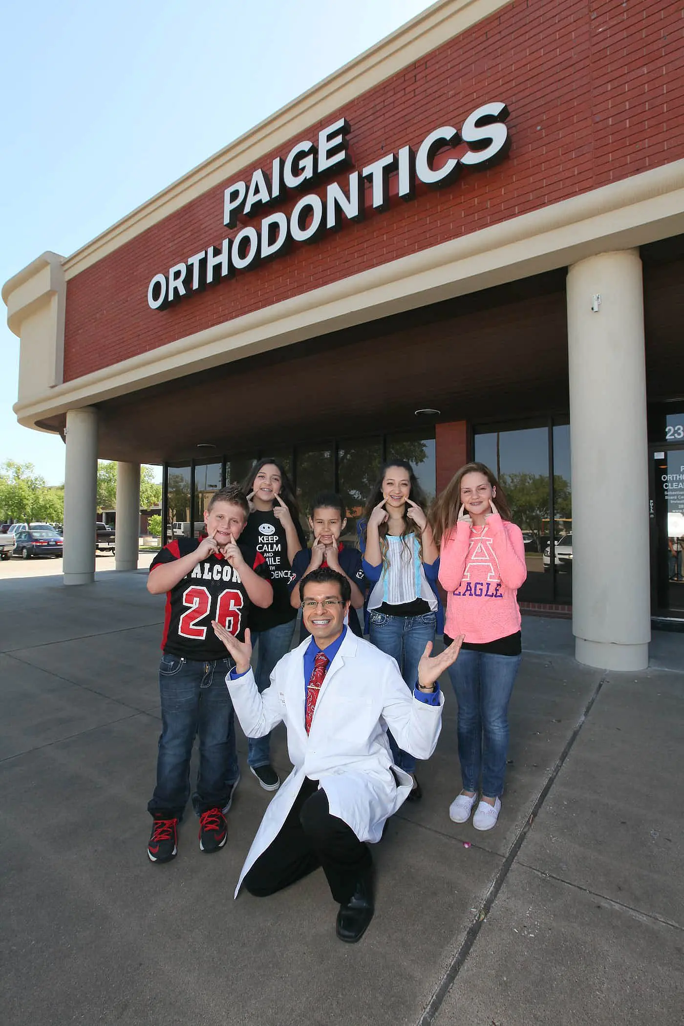 Kneeling in front of five smiling children, a dentist in a white coat poses outside the building marked "Paige Orthodontics" in Kingwood TX.