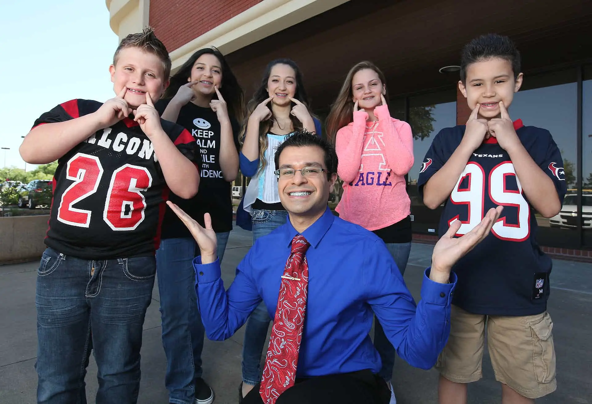 Dr. Sebastian Paige in a blue shirt and red tie, as an orthodontist from Kingwood, poses in front of six smiling children standing outside a TX building.