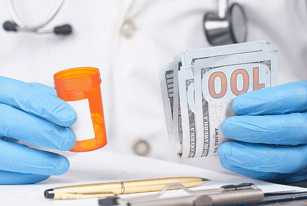 A person in a lab coat and blue gloves holds a small pill bottle in one hand and a stack of hundred-dollar bills in the other to pay for Invisalign service from patien to Paige Orthodontics in Kingwood, TX