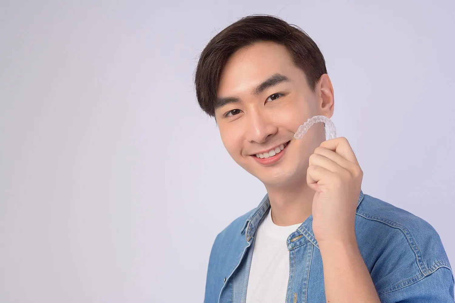 Wearing a denim shirt, a person from Kingwood TX smiles while holding a clear dental aligner against a light gray background.