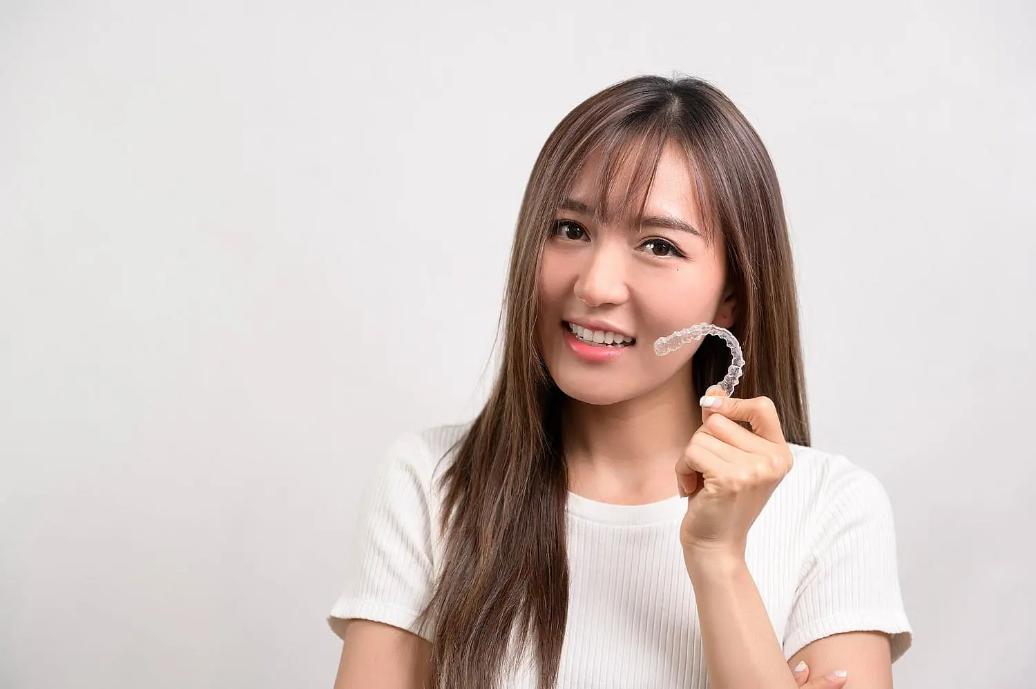 a woman in a white shirt smiles at the camera while holding up a clear dental aligner at Paige Orthodontics in Kingwood, TX