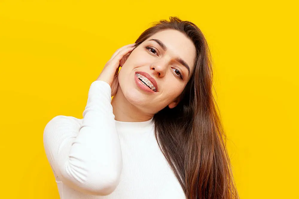 In front of a bright yellow background, a woman with long hair and clear ceramic braces braces, wearing a white shirt, smiles in Kingwood TX.