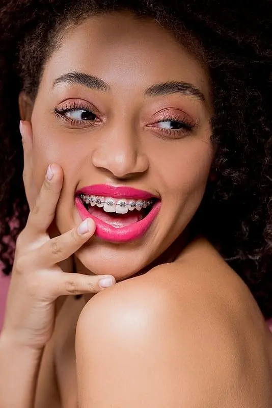 A smiling woman with curly hair and pink lipstick showing metal braces on teeth at Paige Orthodontics in Kingwood, TX